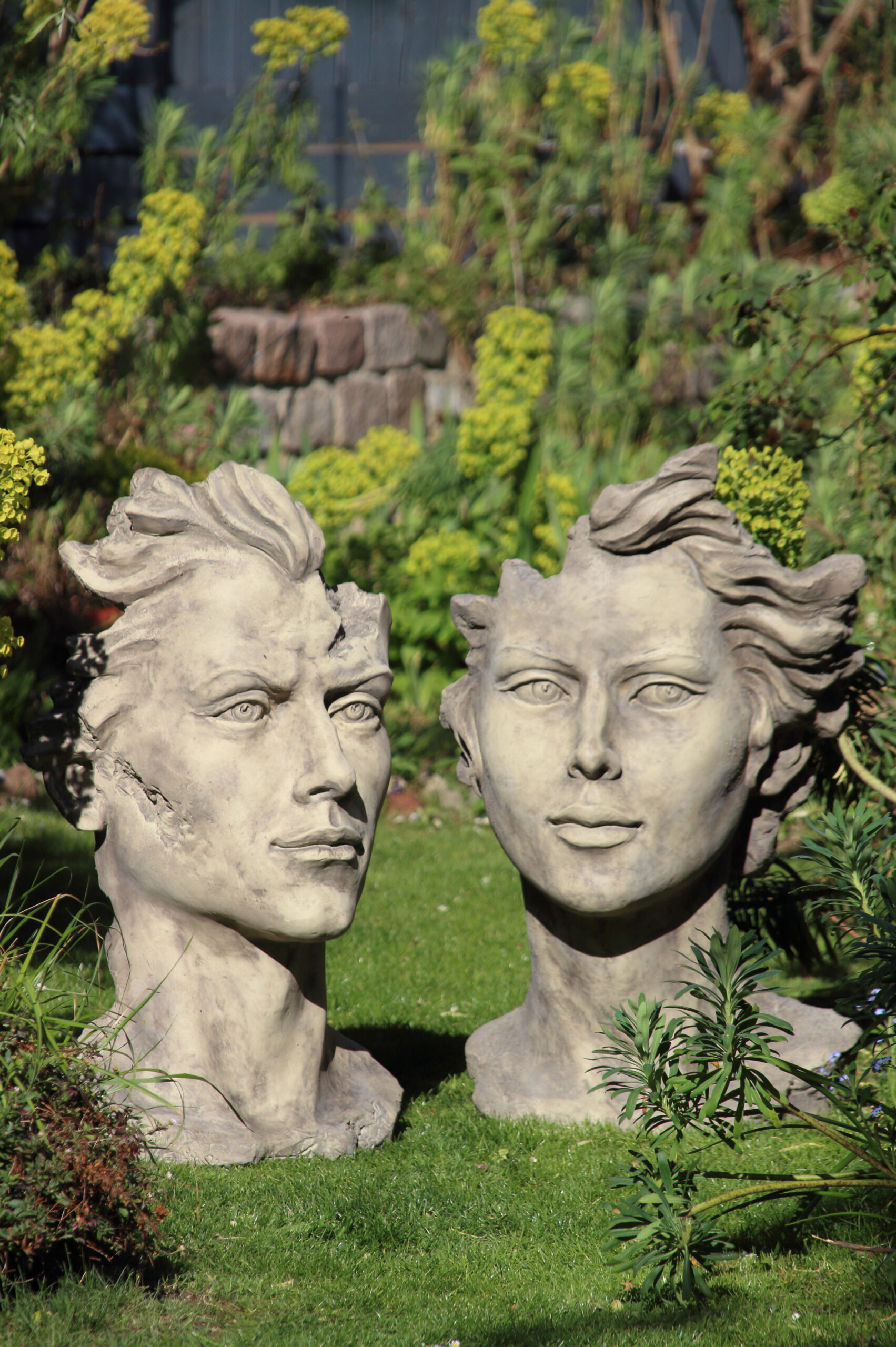 Catálogo 2022 Imagen de uso con las estatuas Liam Cara y Emma Cara, bustos de estilo moderno colocados en un jardín con césped y plantas.