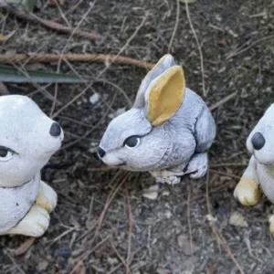 Imagen de contexto: Colección de figuras de conejos Cele, Mele y Pele (familia), de piedra artificial, decorando un jardín con tierra y vegetación.