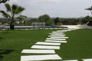 Camino curvo formado por placas decorativas de hormigón de color blanco sobre césped artificial en un jardín exterior con piscina y palmera.